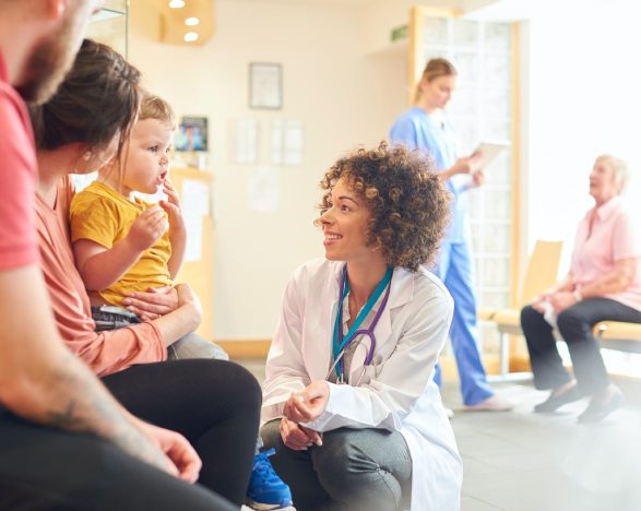 A female doctor talks to a mother and father with a toddler in a waiting room. A nurse talks to another patient in the background.
