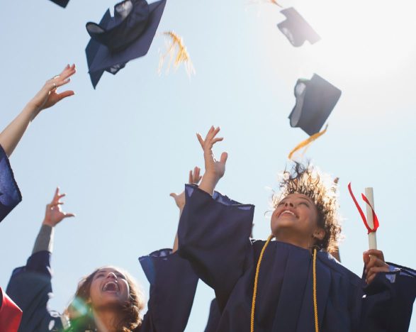 Happy and excited graduates in graduation gowns toss their caps in the air.