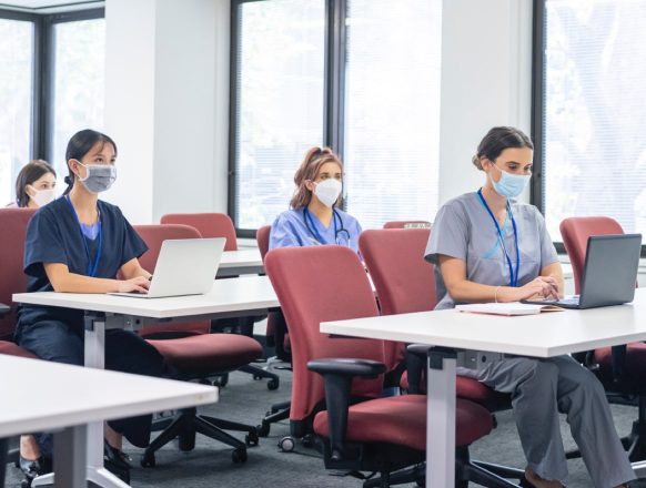 Nurses wearing facemasks are sitting at tables in a classroom working on laptop computers.