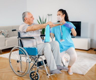 A female nurse kneels next to an older male patient in a wheel chair.